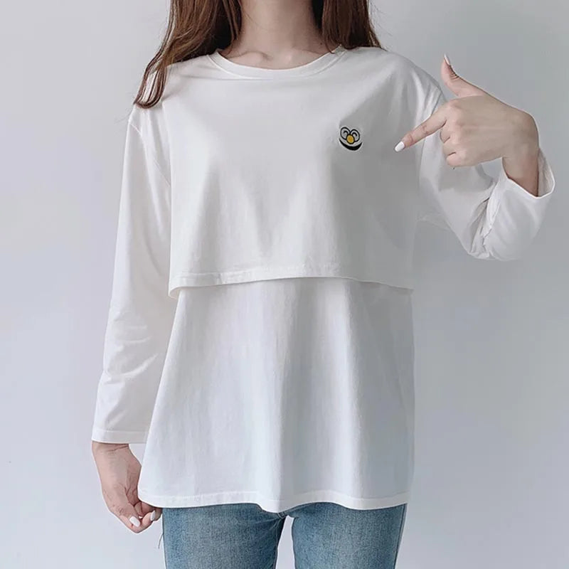 Person wearing a white long-sleeve shirt with a small design on the chest, pointing to it.